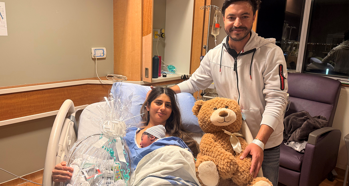 In a hospital room two parents, one holding a newborn in a hospital bed on the left and one standing to the right of the hospital bed holding a teddy bear toy.