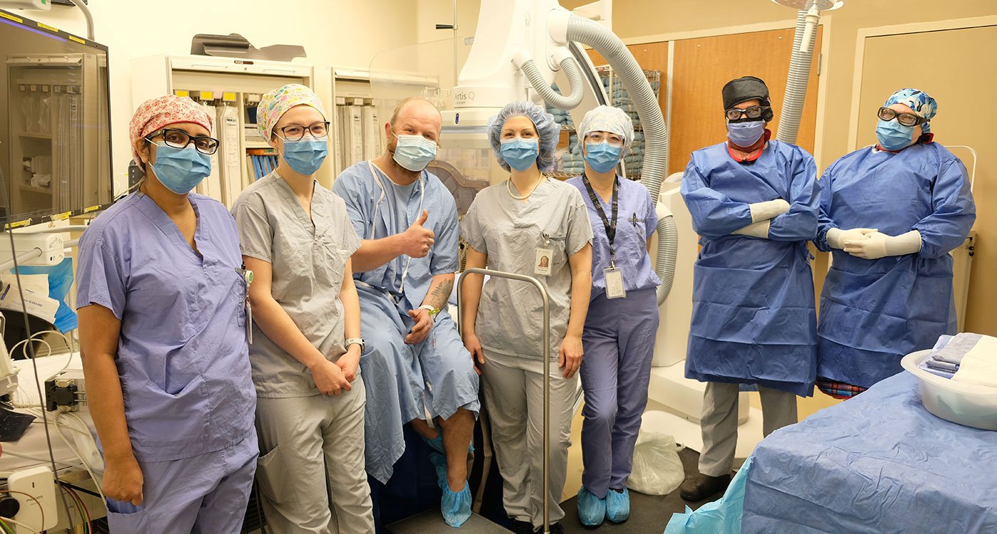 Patient Ryan Siddle (sitting on the table) is surrounded by members of Osler's Cardiology team in an operating room