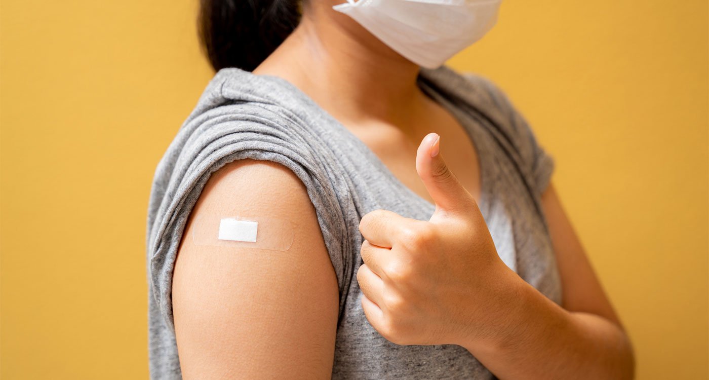 Masked women gives thumbs up while showing bandage on arm after receiving a needle