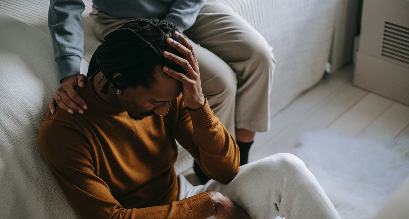 Man sitting on the floor holding his head while a loved one comforts him