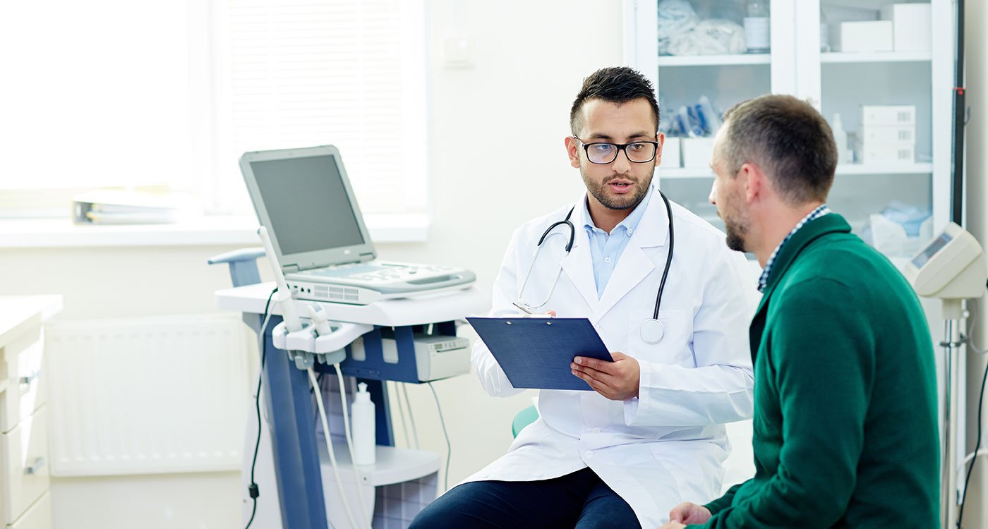 A male physician speaks with his male patient in a clinical setting