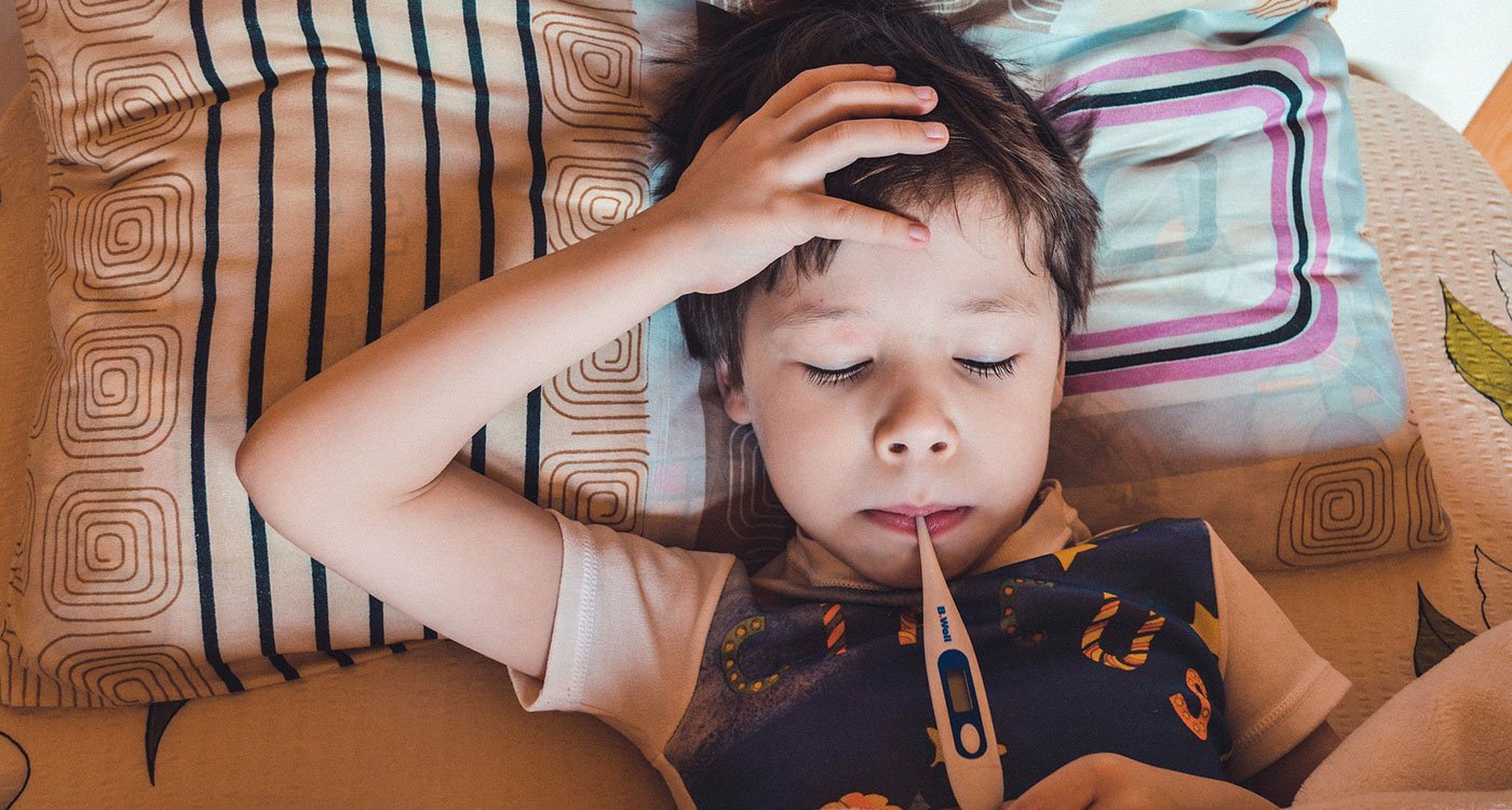 Little boy in bed holds his head with thermometer in his mouth